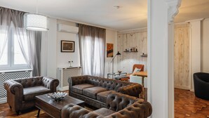A living room with a brown leather sofa, a wooden coffee table, and a white console table. There is a window with curtains, a wall-mounted air conditioner, and a painting.