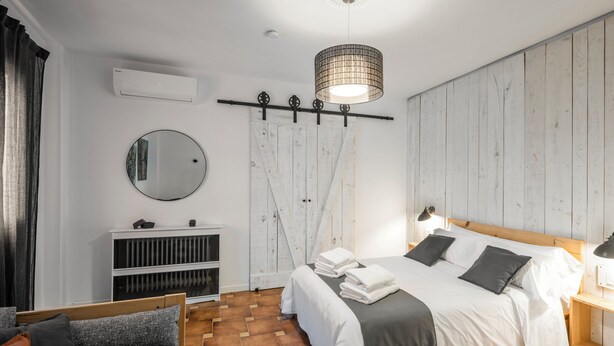A modern bedroom with a wooden floor, a large bed with white and gray bedding, a bedside table with a lamp, a wardrobe, a round mirror, and an air conditioning unit.