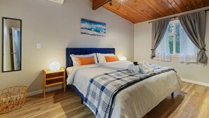 Un dormitorio con techo de madera, una cama con cabecera azul, una mesita de noche con lámpara, un aire acondicionado de pared y una ventana con cortinas.