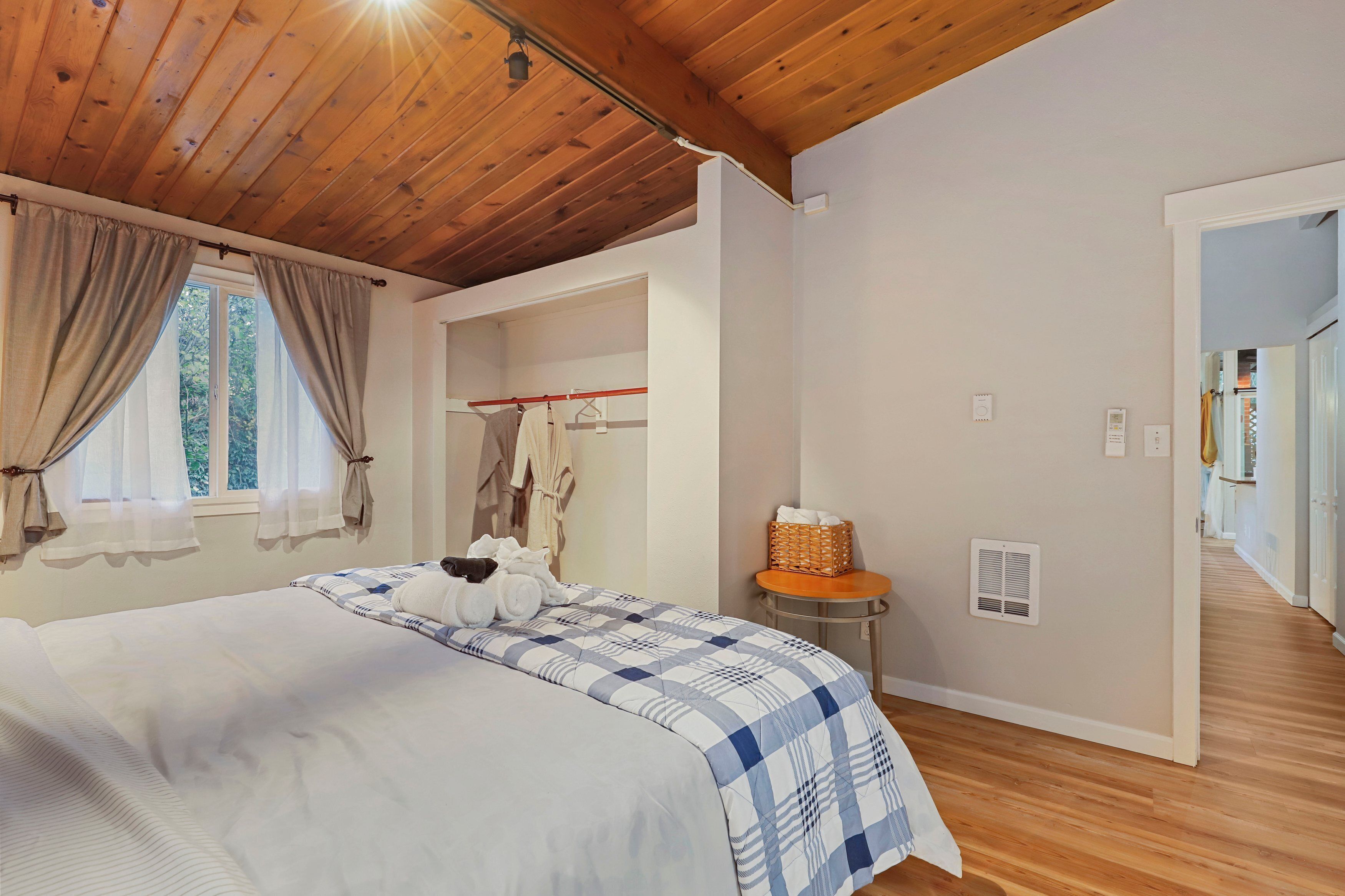 Une chambre avec un plafond en bois, un lit recouvert d’une courtepointe à carreaux bleus et blancs, une table de chevet avec un panier, et une fenêtre avec des rideaux.