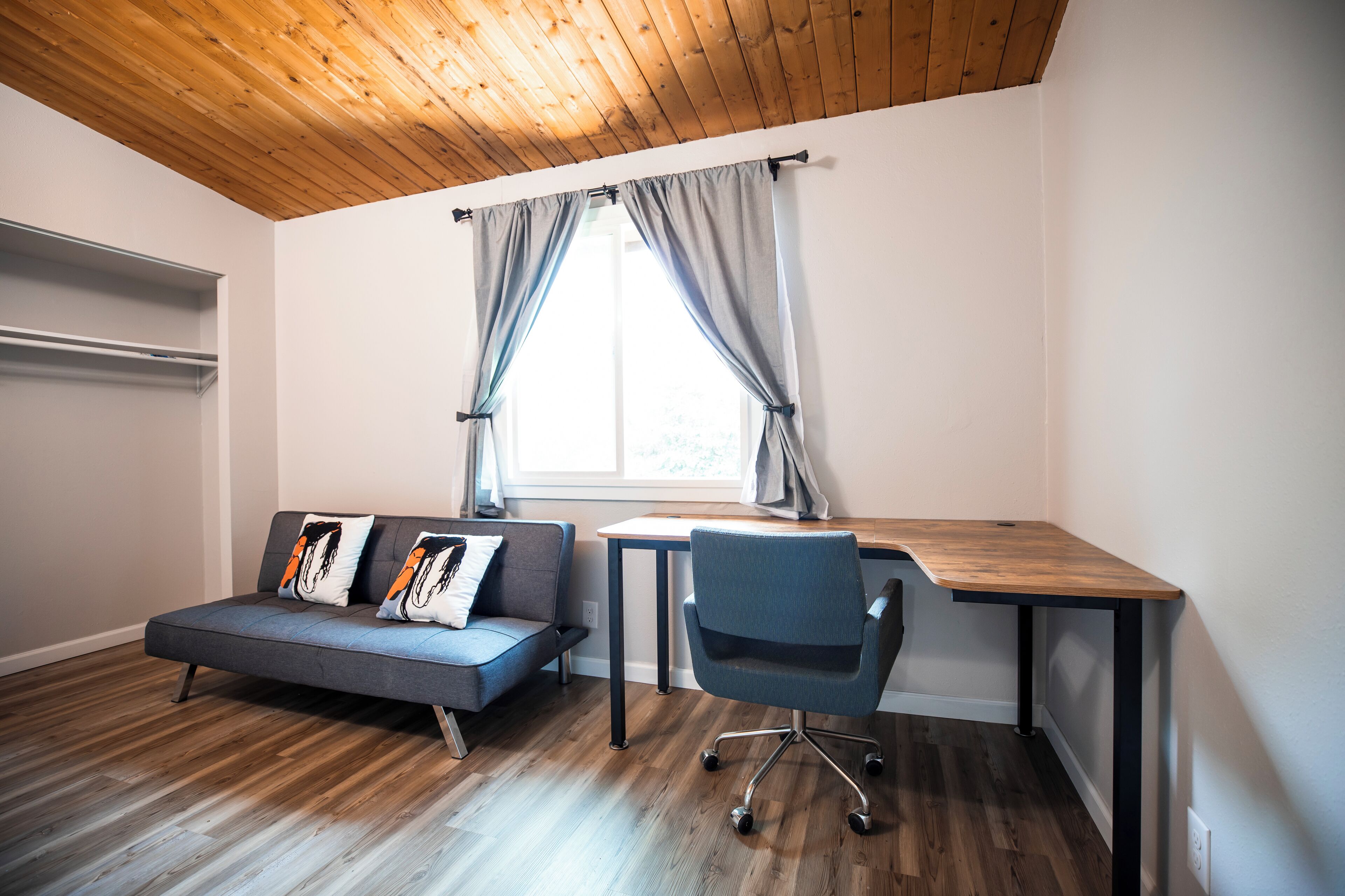 Una habitación moderna con techo de madera, un sofá gris con cojines decorativos, una silla de oficina azul y un escritorio de madera.