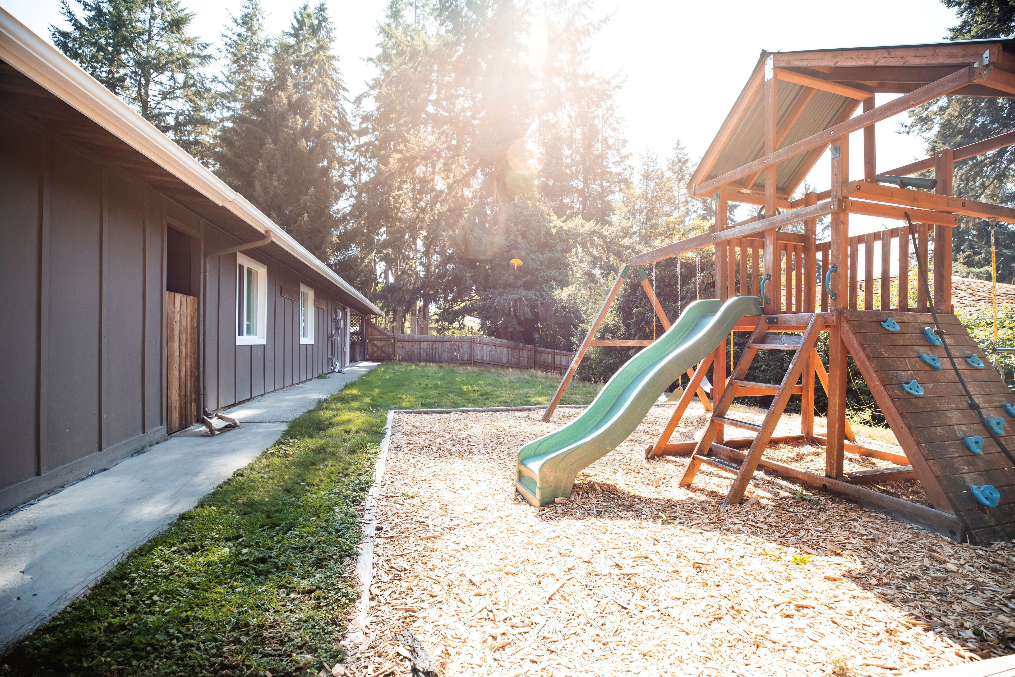Ein Spielplatz mit Rutsche und Kletterwand, umgeben von einer Holzspielkonstruktion und einem Gebäude mit Fenstern.