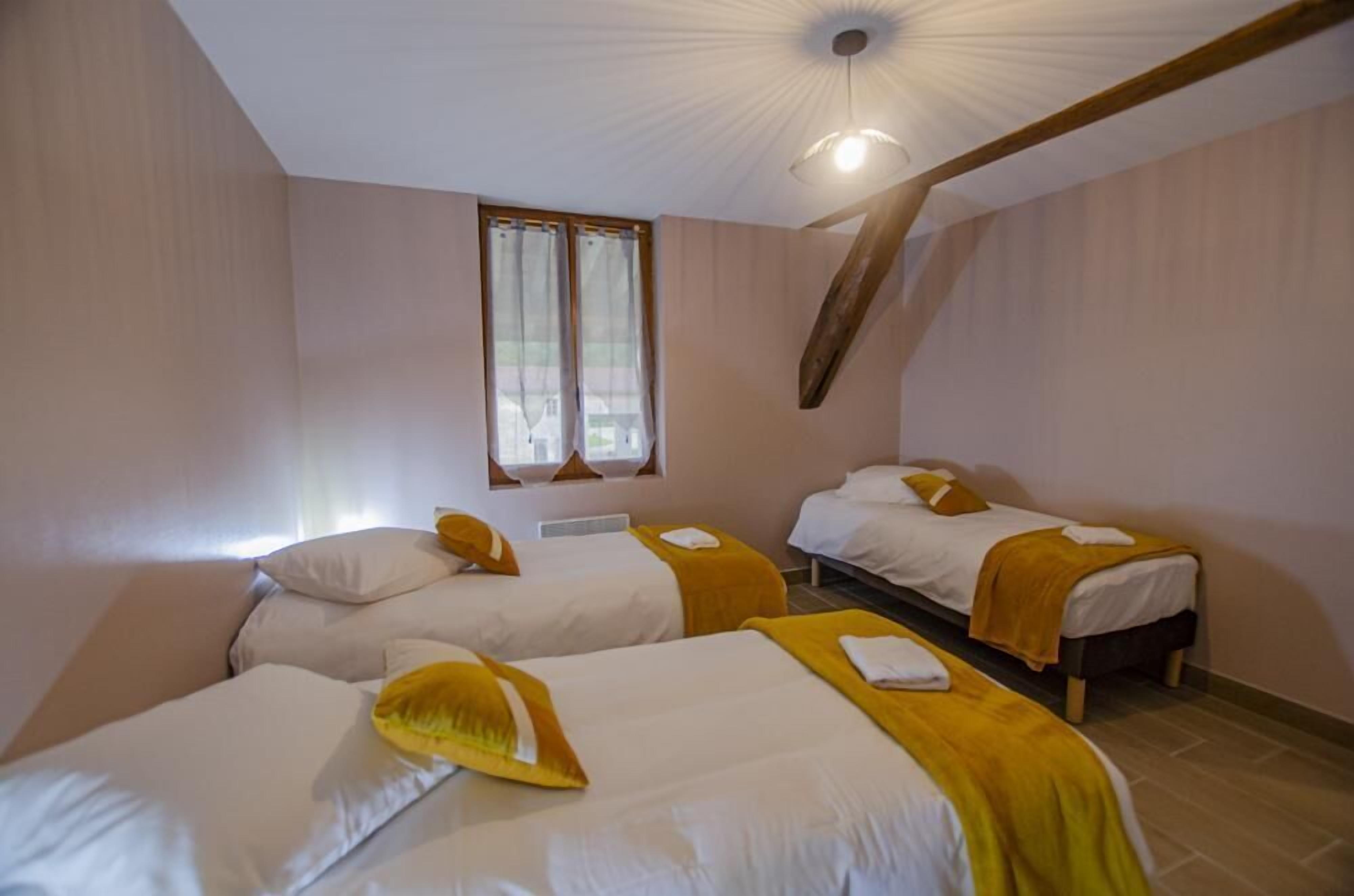 A room with three beds, each with a white mattress and a yellow blanket. There is a window with curtains, a wooden beam on the ceiling, and a light fixture.