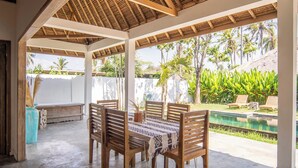 A covered outdoor dining area with a pool view, wooden chairs, and a table with a patterned tablecloth.