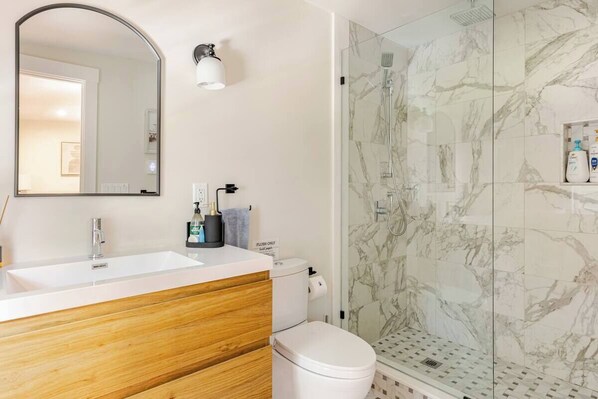 A modern bathroom with a white sink, wooden vanity, a toilet, and a glass shower enclosure with marble tiles. - Beautiful cottage, Close to Wychmere & Beach, Family relax, king bed & fire pit! (Harwich)