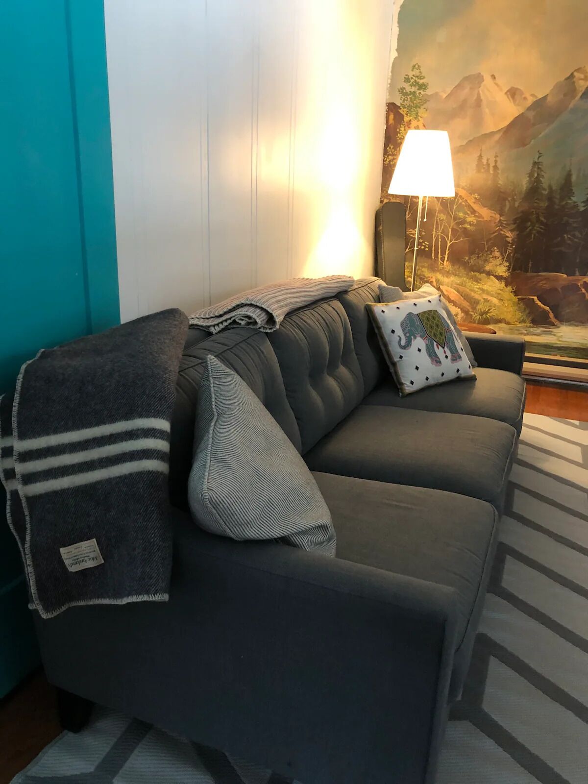 A dark grey sofa with a blanket and pillows, a lamp, and a mountainous mural in the background.