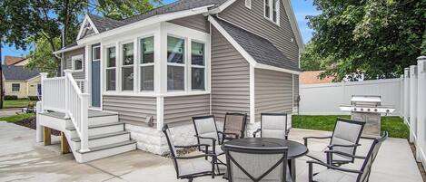 A house with a covered patio area featuring a table and chairs, a grill, and a small deck with steps.