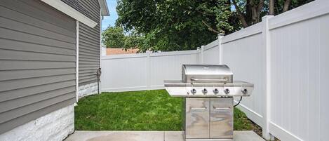 Una parrilla de acero inoxidable en un patio trasero con cerca blanca y una casa al fondo.