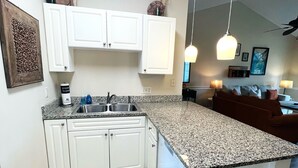 Cocina con armarios blancos, encimera de granito y pileta de acero inoxidable. Se observan luces colgantes y un ventilador de techo.