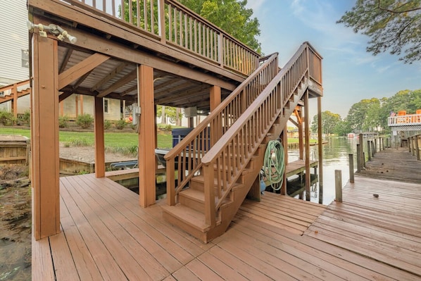 Una terraza de madera con una escalera que lleva a un área cubierta, un muelle que se adentra en el agua y una pequeña embarcación amarrada cerca.