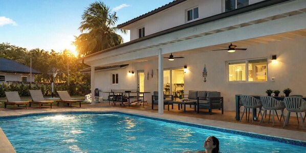 Una piscina con sillas de descanso y mesas, un patio cubierto y un edificio con ventanas y ventilador de techo.