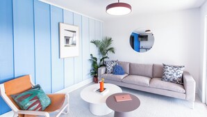 Un salon moderne aux couleurs bleu et blanc, agrémenté d’un miroir rond, d’un canapé avec des coussins à motifs, d’une table basse et d’un fauteuil avec un coussin coloré.