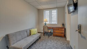 A compact room with a grey sofa, a wooden desk, a chair, a lamp, and a television mounted on the wall. - Cozy Suite in a Revamped Dorm Room! (Nashville)