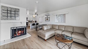 Ein modernes Wohnzimmer mit Kamin, einem beigen Sofa, einem Holztisch und einem Essbereich, im Hintergrund eine Küche.