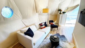 A modern living room with a white sofa, a small black side table, a floor lamp, and a view through a tent-like structure.