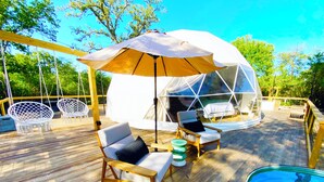 A wooden deck with a glass igloo structure, white cushioned chairs, and a pool.