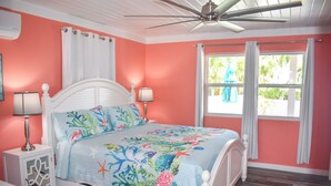 A bedroom with a bed, white nightstands, a ceiling fan, a window with curtains, and a view of greenery outside. - Lillian's Downstairs - Island Comfort & Charm (Green Turtle Cay)