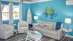 A living room with a blue wall, a white sofa, a glass coffee table, two white armchairs, a small side table with a lamp, and a decorative crab on the wall. - Lillian's Downstairs - Island Comfort & Charm (Green Turtle Cay)