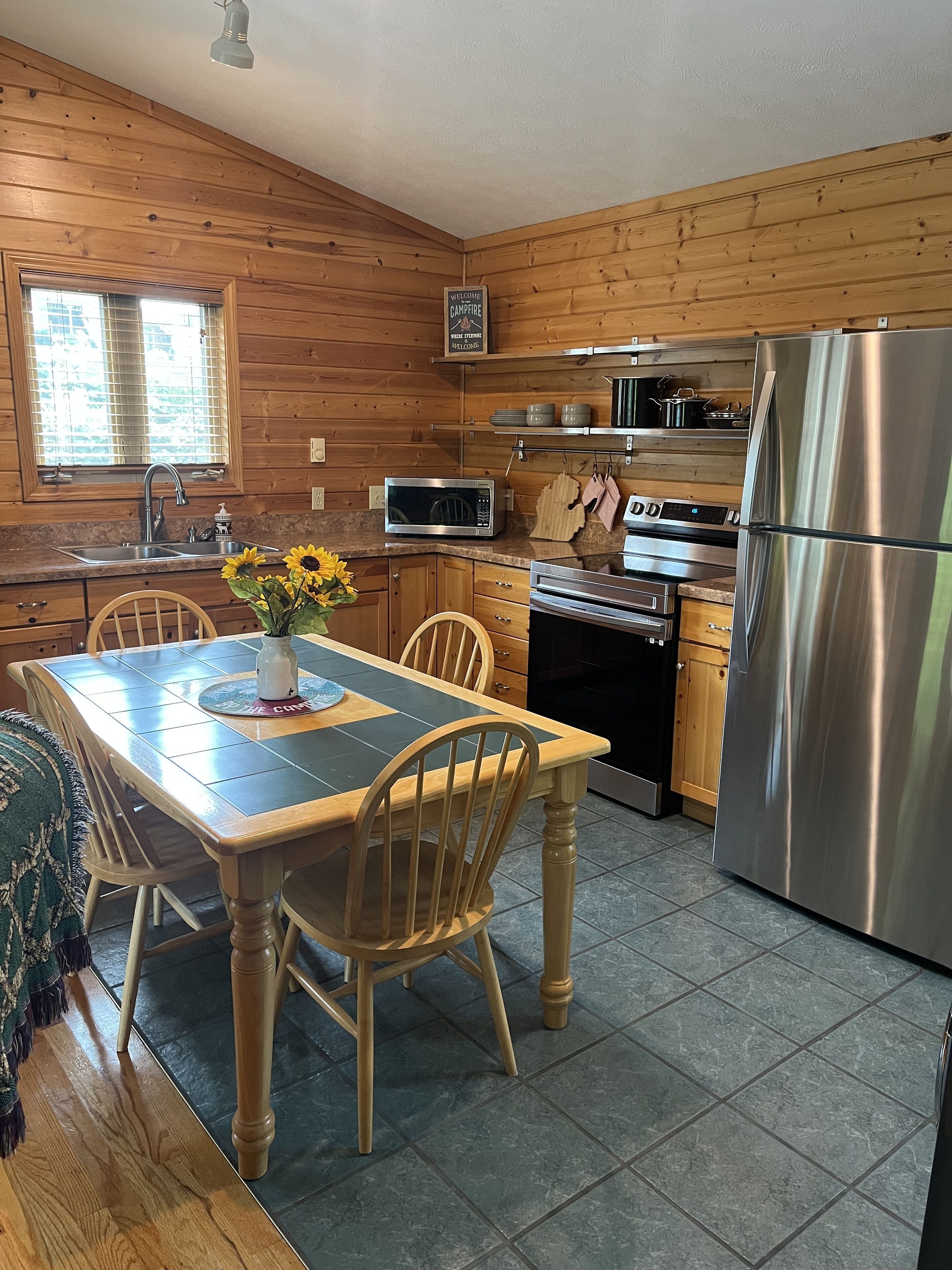 A cabin-style kitchen with wooden walls, a dining table with chairs, a refrigerator, a microwave, and a sink.
