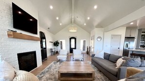 A spacious living room with a fireplace, a large TV mounted on a brick wall, and a modern kitchen in the background.