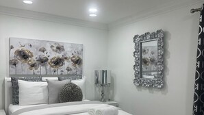 A bedroom with a white bed, a decorative wall piece, a mirror, and a nightstand. - Do you want to select a perfect place to know Miami?. This is R and R Hostal.  (Miami)