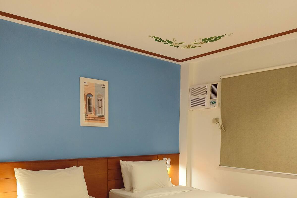 A hotel room with two wooden beds, a nightstand, a wall-mounted air conditioner, and a framed picture on the blue wall.