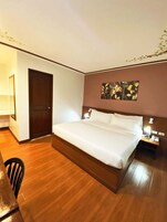 A hotel room with a large bed, a wooden floor, a desk with a chair, a nightstand with a lamp, and a door.