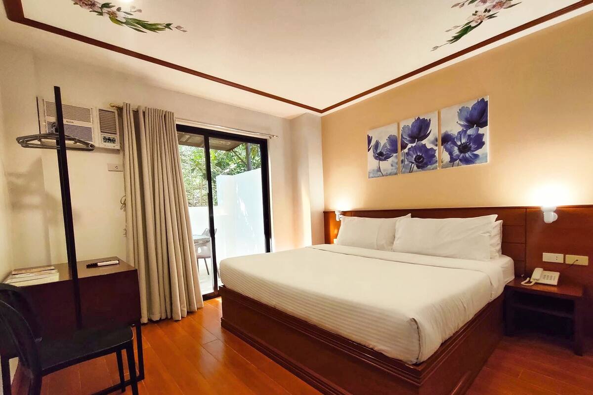 A hotel room with a large bed, a desk with a chair, a telephone, and a painting of flowers on the wall.