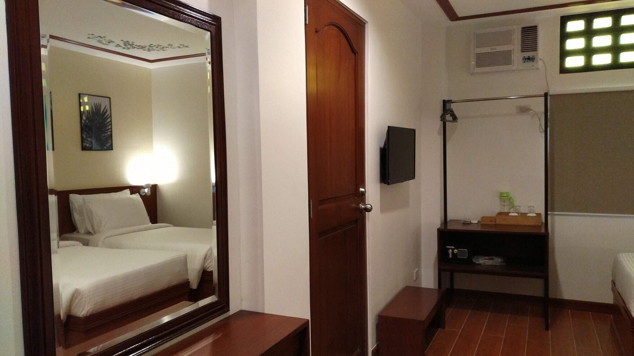 A hotel room with a bed, a mirror, a door, a shelf with toiletries, and a window with blinds.