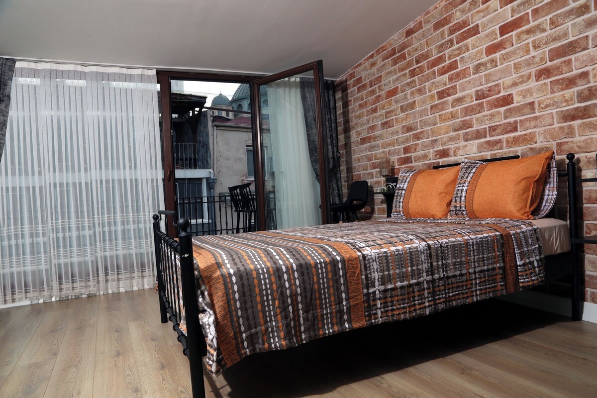 Ein Schlafzimmer mit einem Bett, einem Fenster mit durchsichtigen Vorhängen, einer Ziegelwand und einem Balkon mit Stühlen.