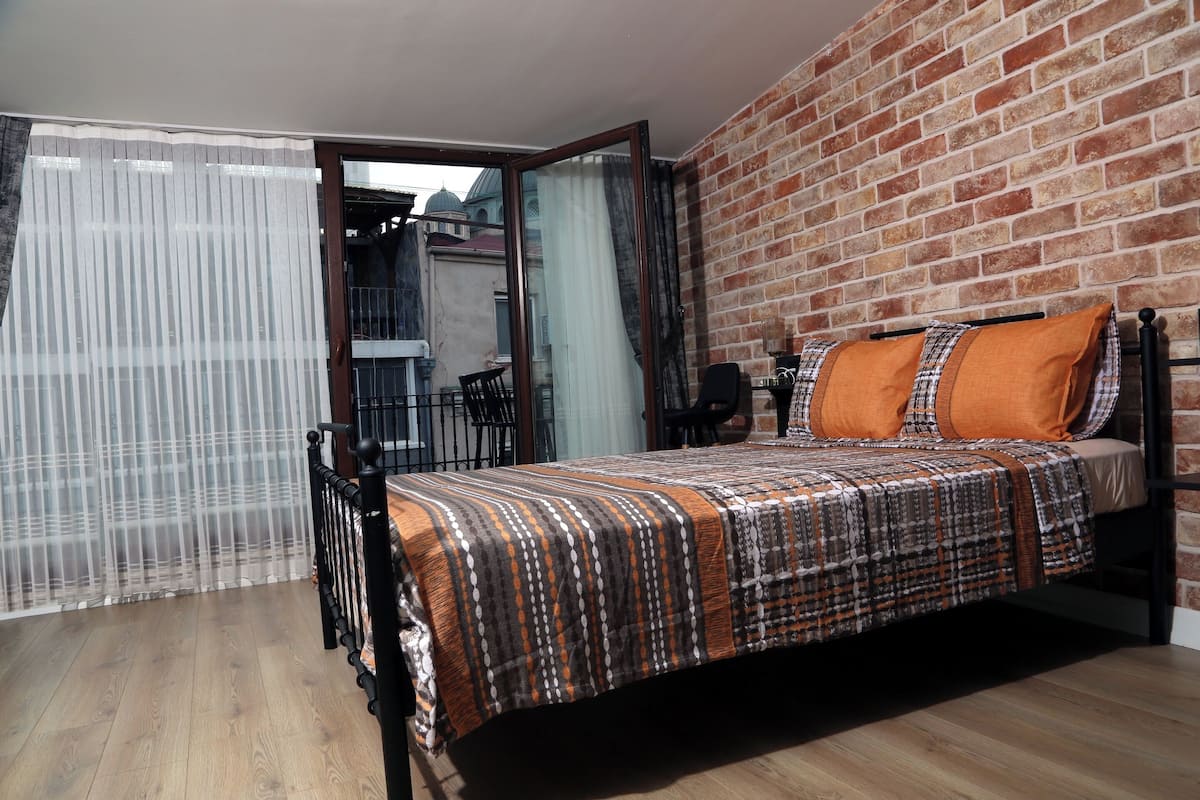 Ein Schlafzimmer mit einem Bett, einem Fenster mit durchsichtigen Vorhängen, einer Ziegelwand und einem Balkon mit Stühlen.