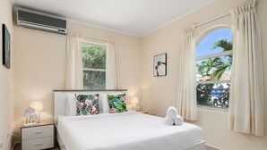 Une chambre à coucher avec un lit, des tables de chevet, un climatiseur, une fenêtre avec des rideaux et une vue sur des palmiers.