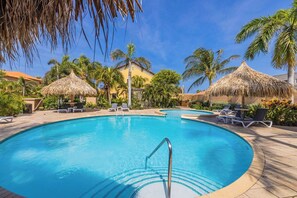 Un área de piscina con sillones de descanso y sombrillas de paja, rodeada de palmeras y un cielo despejado.