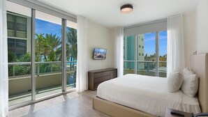 A modern hotel room with a large bed, a desk, a chair, a TV, and a balcony with a view of palm trees and buildings. - Ocean View Wraparound Balcony and Rooftop Pool-CS1 (Hollywood)