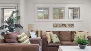 Ein Wohnzimmer mit einem braunen Sofa, einem weißen Couchtisch und einem Schild über den Fenstern mit der Aufschrift 'GATHERING ROOM'.