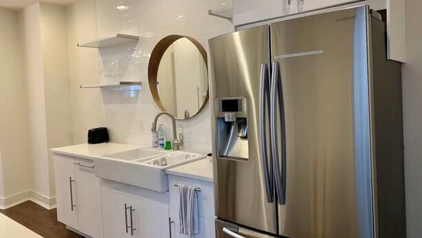 Eine moderne Küche mit weißen Schränken, einem Edelstahlkühlschrank, einem eingebauten Backofen und einem großen runden Spiegel über der Spüle.