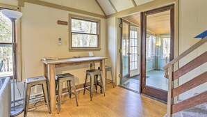 Una habitación con un escritorio de madera, dos taburetes, una ventana y una puerta que da a otra habitación.