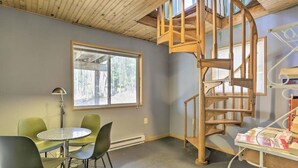 Una habitación con una escalera de caracol, una mesa redonda con sillas, un ventanal y una estantería blanca con libros.