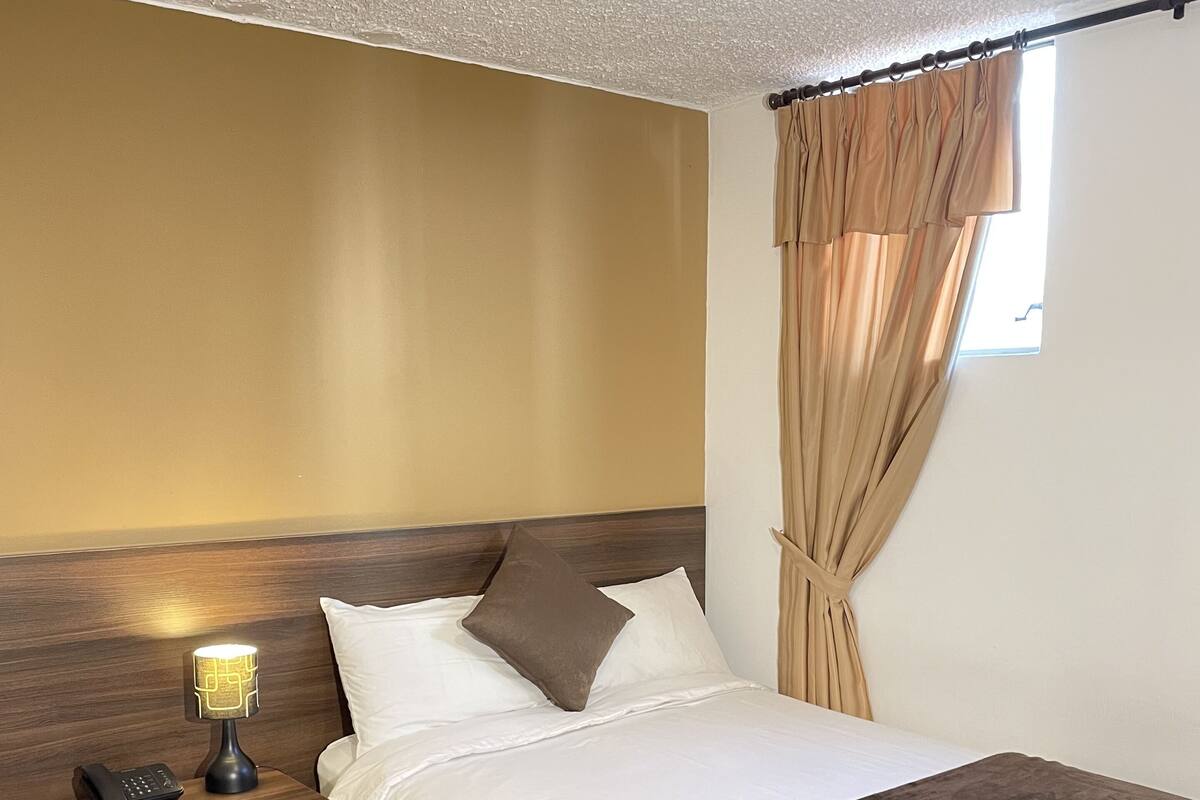 A hotel room with a bed, a bedside table with a lamp, a telephone, and a window with curtains.