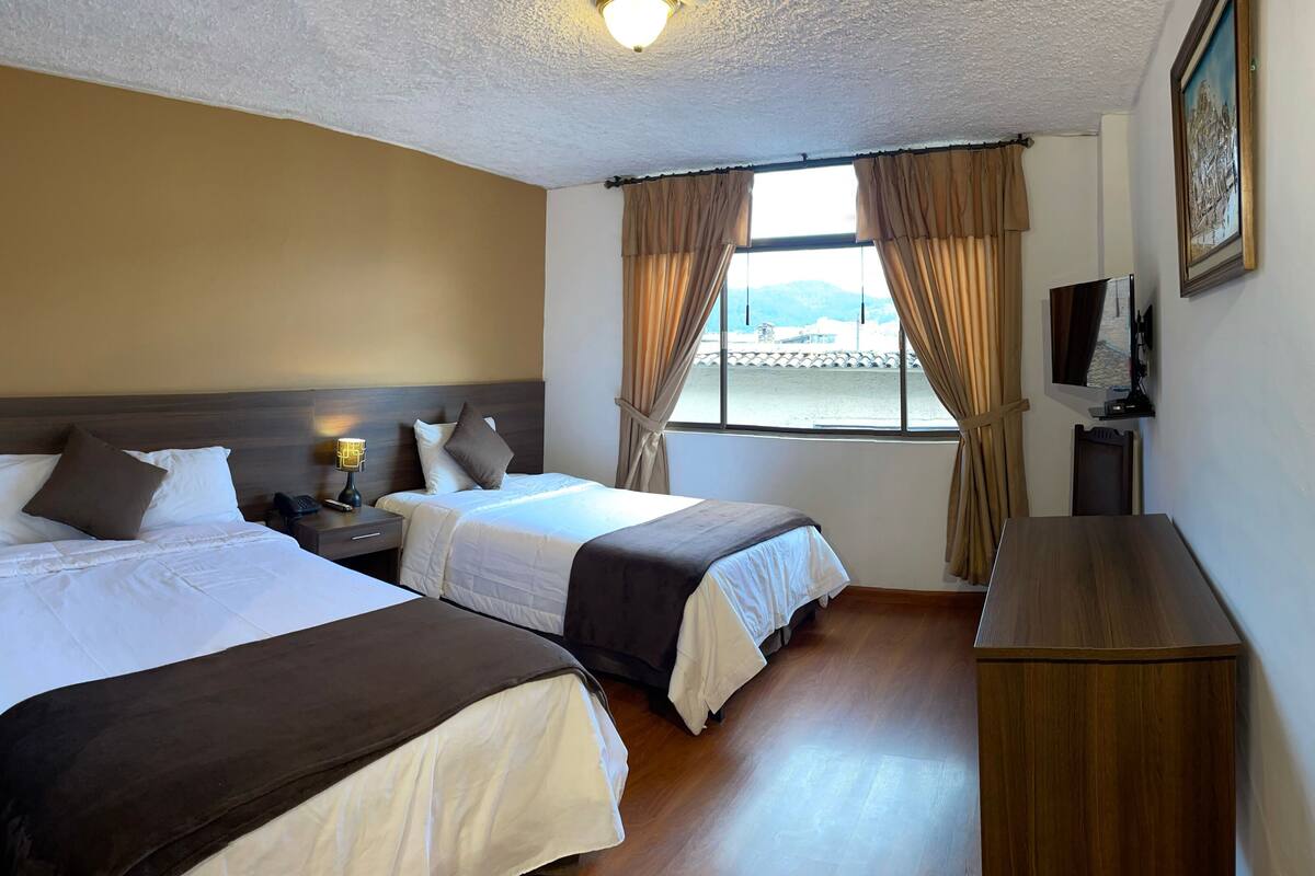 A hotel room with two beds, a wooden nightstand, a desk, a chair, a TV, and a window with curtains.