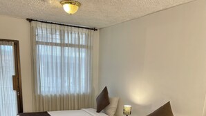 Quarto de hotel com duas camas, cada uma com colcha branca e castanha, luminária de cabeceira e janela com cortinas leves.