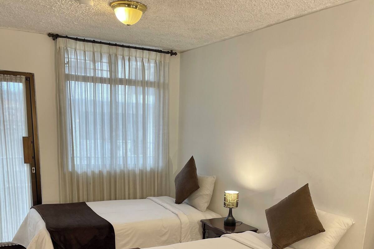 A hotel room with two beds, each with a white and brown bedspread, a bedside lamp, and a window with sheer curtains.
