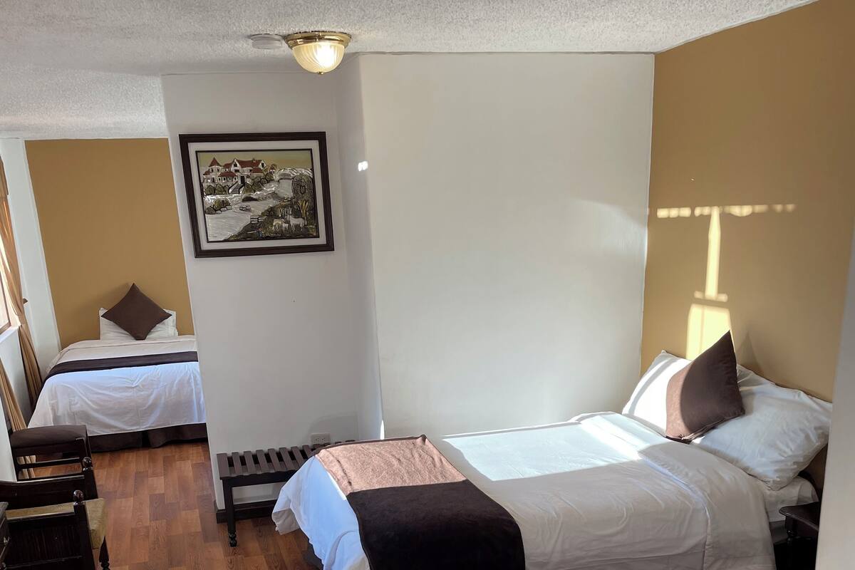 A hotel room with two beds, wooden flooring, a framed picture on the wall, and a bedside table.