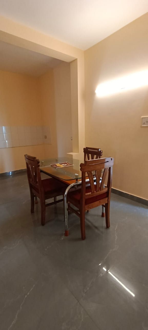 Uma sala de jantar com mesa de vidro e cadeiras de madeira, piso ladrilhado e luminária fixada na parede.