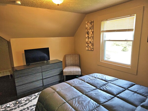 Un dormitorio con una cama, una silla, un tocador, un televisor y una ventana con persianas.