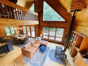 Un intérieur de chalet chaleureux avec une chambre en mezzanine, un coin repas et un salon doté d’une grande fenêtre donnant sur l’extérieur.