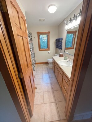 Une salle de bain avec des meubles en bois, un lavabo blanc, des toilettes et une fenêtre donnant sur les arbres.