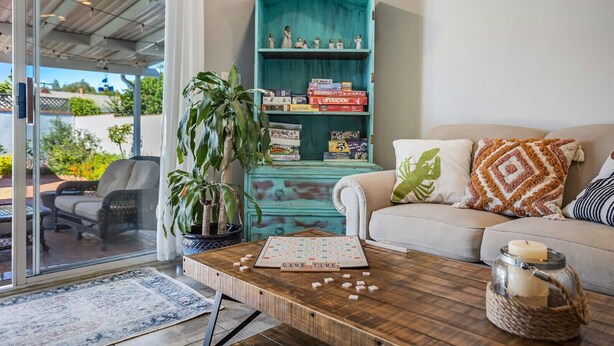 A cozy living room with a teal bookshelf, a coffee table with board games, a potted plant, and a sofa with decorative pillows. - Relax at this Family Home w/ Solar heated Pool & Game Room. Close to everything! (San Diego)