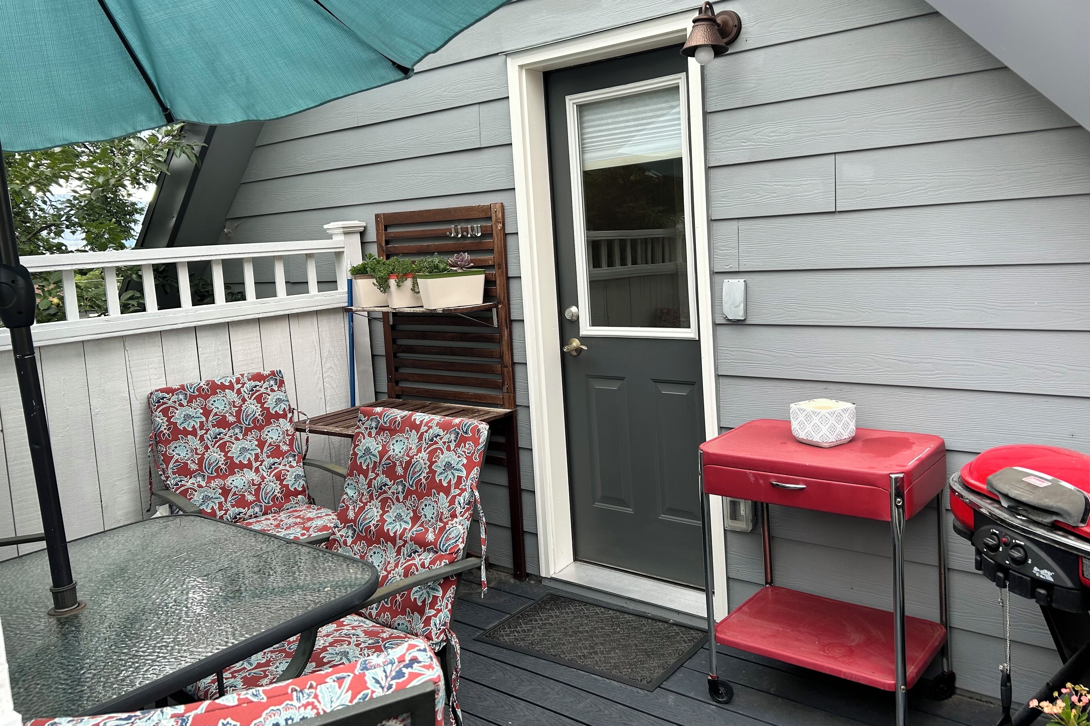 Een terras met een glazen tafel en twee gestreept beklede stoelen, een rode barbecue kar en een deur met een raam.