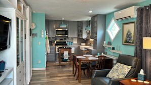 A modern kitchen with stainless steel appliances, a dining area with a wooden table and chairs, and a living room with a leather sofa and a TV.